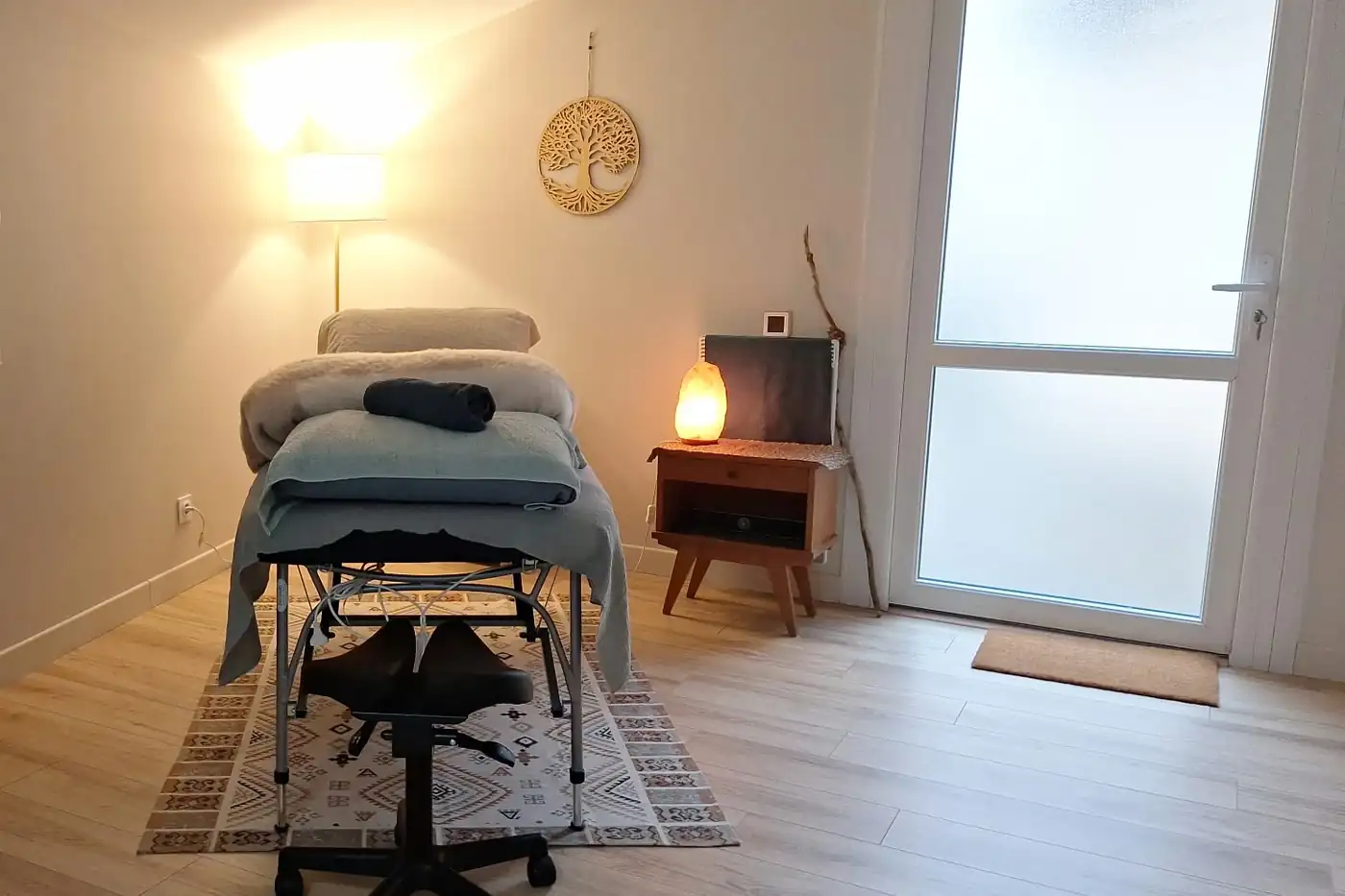 Une pièce claire et chaleureuse : une table de réflexologie recouverte de plaids, un lampadaire, un arbre en bois découpé au mur et une porte vitrée.